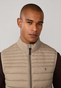 Chaleco acolchado beige con cuello alto, con costuras horizontales y cierre de cremallera. Detalle de logo sutil en el pecho izquierdo, combinado con un suéter oscuro.