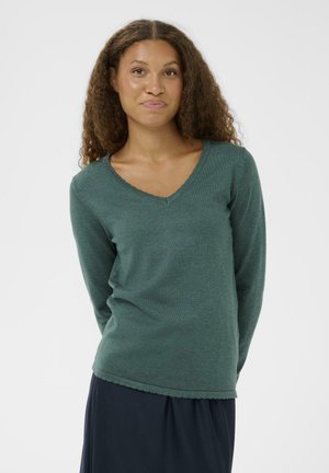 Mujer con cabello rizado, vistiendo un suéter de cuello en V de manga larga en color teal y una falda oscura, de pie frente a un fondo blanco liso.