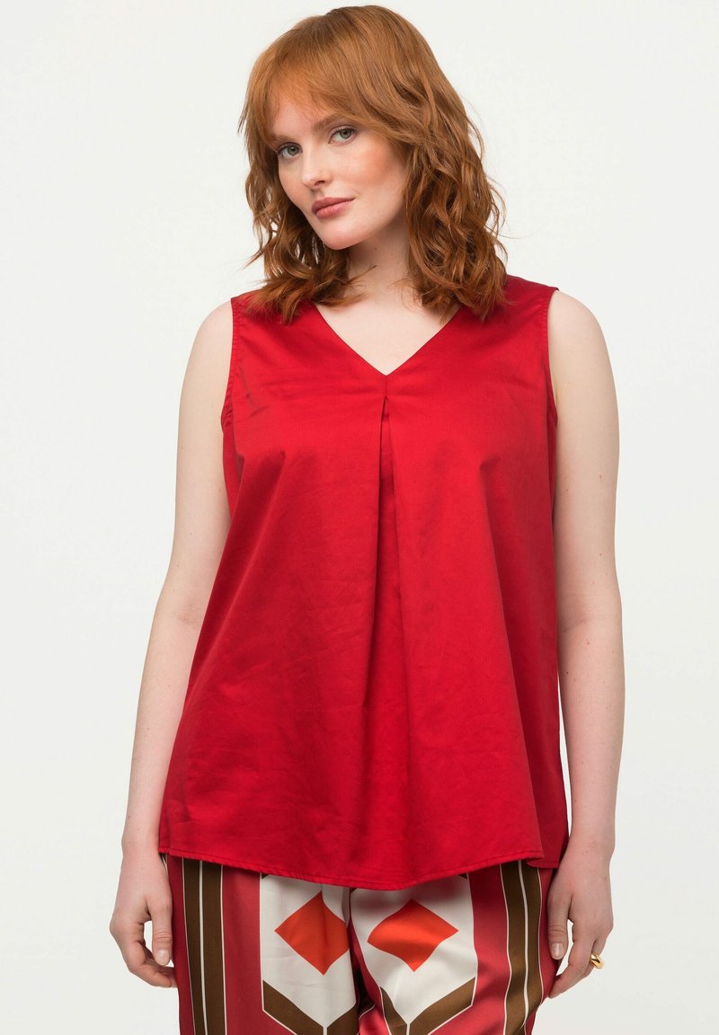 Top rosso senza maniche con scollatura a V e pieghe decorative. Tessuto morbido, vestibilità rilassata, indossato con pantaloni a palazzo a fantasia.