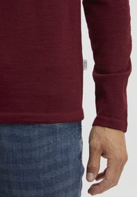 Burgunderfarget langermet skjorte laget av ribbestrikket stoff, med avrundet kant og en liten etikett på siden. Brukt sammen med blå denimjeans.