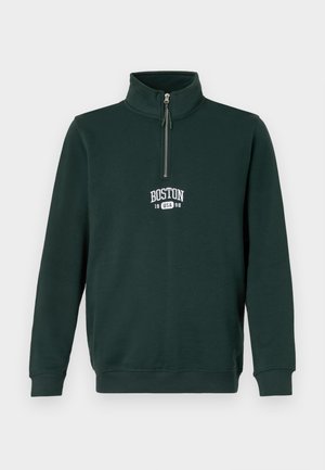 Mørkegrønn genser med kvart glidelås. Har "BOSTON 1998" i hvit skrift på brystet. Laget av mykt, teksturert stoff.