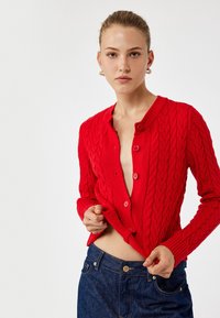 Roter gestrickter Cardigan mit Zopfstrickmuster, runder Halsauschnitt und Knopfleiste. Kombiniert mit dunkelblauen, hochgeschnittenen Jeans.