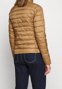 Brun dynejakke med horisontalt quiltet design, høj krave og en glat, skinnende finish. Kombineret med mørkeblå jeans.