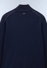 Pull bleu marine à col montant, petit détail rayé rouge, blanc et noir sous le col et coutures d’épaules légèrement visibles.