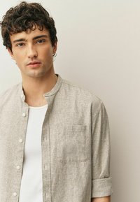 Camicia di lino grigio chiaro con colletto alla coreana, bottoni frontali e una tasca sul petto; indossata sopra una maglietta bianca. Tessuto liscio.