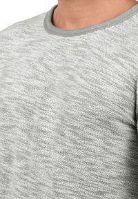 Pullover en tricot gris clair texturé avec un motif subtil, dotée d'un col rond côtelé gris contrastant. Le tissu semble doux et tissé.