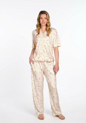 Frau trägt cremefarbenes, florales Kurzarmoberteil und passende lockere Hose mit Taschen, steht mit einer Hand in der Tasche und Sandalen.