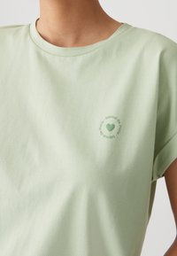 T-shirt di cotone verde chiaro con scollo rotondo e maniche corte, decorata con un logo circolare stampato di colore verde sul petto.