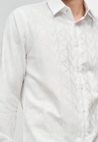 Κοντινό πλάνο ενός ατόμου που φοράει λευκό πουκάμισο με κουμπιά και διακριτικό γεωμετρικό μοτίβο, με ανοικτό γιακά.