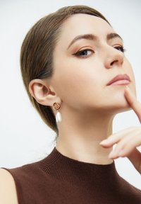 Pendientes de aro dorados con un colgante de gota perlado. La modelo lleva un suéter de cuello alto marrón, mostrando el diseño de la joyería.