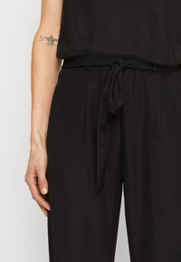 Mono de tela negra con un corte relajado, que presenta una cintura atada y piernas anchas. Textura suave con detalles de diseño mínimos, sin patrones visibles.