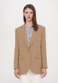 Tan blazer dengan bahu yang terstruktur, lapel bergerigi, dan dua kantong depan. Dipadukan dengan atasan bermotif. Tekstur halus, potongan rapi.