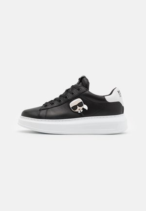 Czarny niskoprofilowy sneaker z białą podeszwą, białą zakładką na pięcie i małą kreskówkową twarzą z okularami przeciwsłonecznymi po boku.