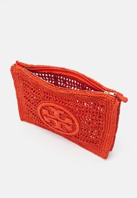 Prostokątna, pomarańczowa torebka na szydełku w kształcie kopertki z otwartym zamkiem błyskawicznym i okrągłym, plecionym emblematem z przodu, wyeksponowana na białym tle.
