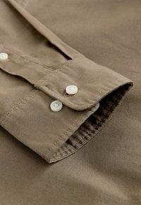 Puño de camisa de algodón marrón con una textura suave, que presenta dos botones de plástico blancos y costuras reforzadas a lo largo de los bordes.