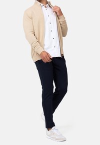 Cardigan beige en tricot avec poignets côtelés, porté sur une chemise blanche  à boutons et un pantalon bleu marine. Complété par des baskets claires.