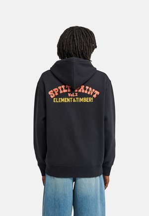 Persoon met dreadlocks die een zwarte hoodie draagt met oranje en gele "Spilt Paint Vol.2 Element & Timber!" tekst op de achterkant, gecombineerd met een blauwe spijkerbroek.