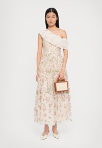 Schouderloze bloemenjurk in lichtroze met groene borduursels, gelaagde tule rok en bijpassende bloemen-sjaal. Bruin vierkant handtasje.