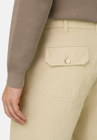 Pantalon beige avec une poche arrière boutonnée porté avec un pull côtelé à manches longues taupe, montrant le bras gauche et la main détendus sur le côté.