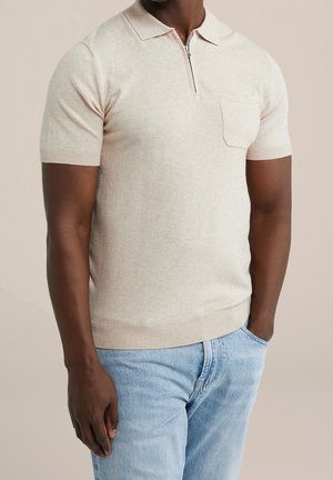 Homme portant un pull en maille beige à manches courtes avec col zippé et poche poitrine, et un jean bleu clair, debout avec une main dans la poche.
