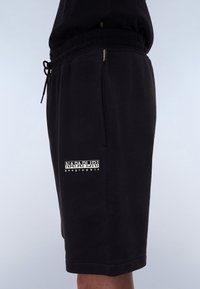Black cotton shorts with an elastic waistband, drawstring closure, and a side logo. Smooth texture and knee-length design.