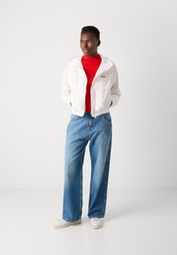 Lightweight white hooded jacket, red high-neck shirt, oversized blue denim jeans, and white sneakers. Notable details include elastic cuffs.