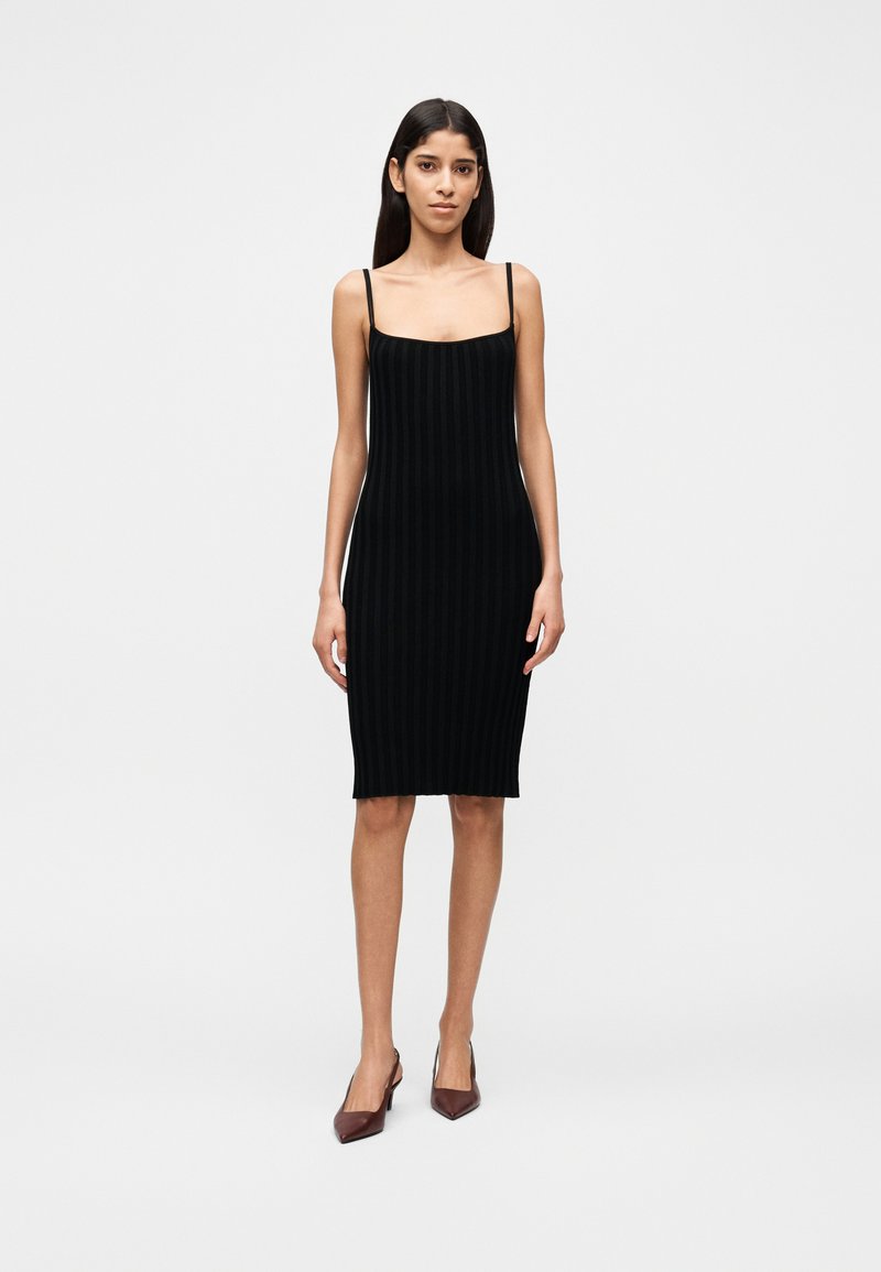 Μαύρο ριμπ πλεκτό φόρεμα με λεπτές τιράντες, εφαρμοστό σχέδιο, μήκος μέχρι το γόνατο, λεία υφή, φορεμένο με καφέ μυτερά παπούτσια με τακούνι.