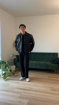 Casaco de couro preto com botões de pressão e detalhe vermelho na manga, usado sobre uma camisa preta e combinado com calças pretas e sapatos brancos.