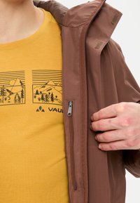 Veste imperméable marron avec fermeture éclair à l'avant, associée à un t-shirt jaune arborant un motif de montagne. Texture lisse, design décontracté.