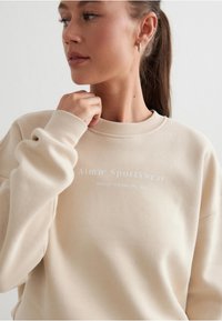 Beige mikina s kulatým výstřihem a vroubkovanými manžetami, na které je v bílém textu napsáno "Aim'n Sportswear". Měkká látka s jednoduchým designem, volný střih.