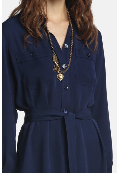 Žena oblečená v námornícky modrom košeľovom šatách s opaskom, s náprsnými vreckami a zlatým reťazovým náhrdelníkom srdcového tvaru a rôznymi príveskami.