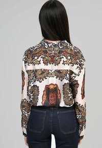 Blouse en soie à manches longues présentant un motif paisley en bleu foncé, rouille et beige sur fond crème clair, avec un design à bouton dans le dos.