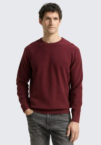 Bordeauxroter gestrickter Pullover mit Rundhalsausschnitt, langen Ärmeln und gerippten Bündchen sowie Saum. Trägt man mit grauen Jeans, die eine glatte Textur aufweisen.