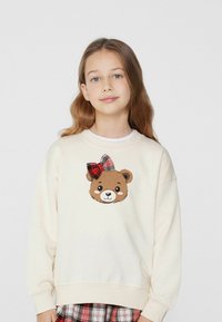Sweatshirt crema con design di faccia d'orso e un fiocco a quadri rosso e nero. Tessuto morbido, vestibilità rilassata e polsini a coste.