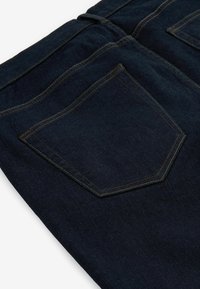 Jeans de mezclilla azul oscuro con una textura suave y costuras doradas, que cuentan con un detalle en el bolsillo trasero en forma hexagonal.