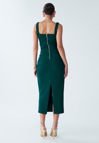 Fitted green dress with a square neckline, back zipper, and ruching detail. Mid-length with a slit at the back, paired with strappy beige heels.