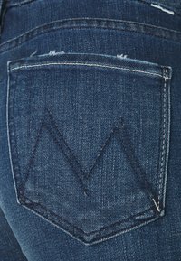 Jeans de denim azul oscuro con un bolsillo trasero que presenta un distintivo patrón de costura en zigzag. El material es texturizado con sutiles detalles de desgaste.