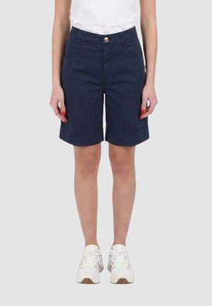 Persona che indossa pantaloncini blu navy fino al ginocchio, scarpe da ginnastica bianche e una canotta bianca, in piedi davanti a uno sfondo grigio chiaro uniforme.