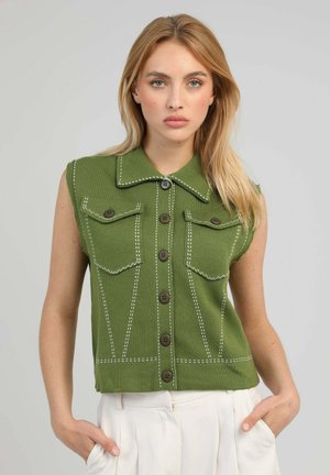 Femme aux cheveux blonds portant un haut vert sans manches boutonné avec des coutures blanches, debout les mains dans les poches devant un fond uni.