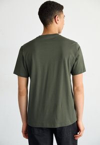 Tricou din bumbac de culoare verde măsliniu, cu guler rotund, mâneci scurte și o textură netedă, vizualizat din spate, având un tiv drept.