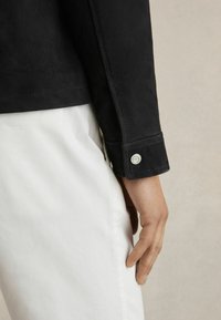 Veste en suède noire avec un gros plan sur le poignet mettant en avant un bouton en métal, associée à un pantalon blanc. Texture lisse, design ajusté.