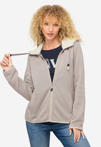 Sudadera con cremallera de color beige claro, con collar forrado de felpa, dos bolsillos frontales y textura acanalada. Tiene una cremallera de contraste y capucha con cordón.