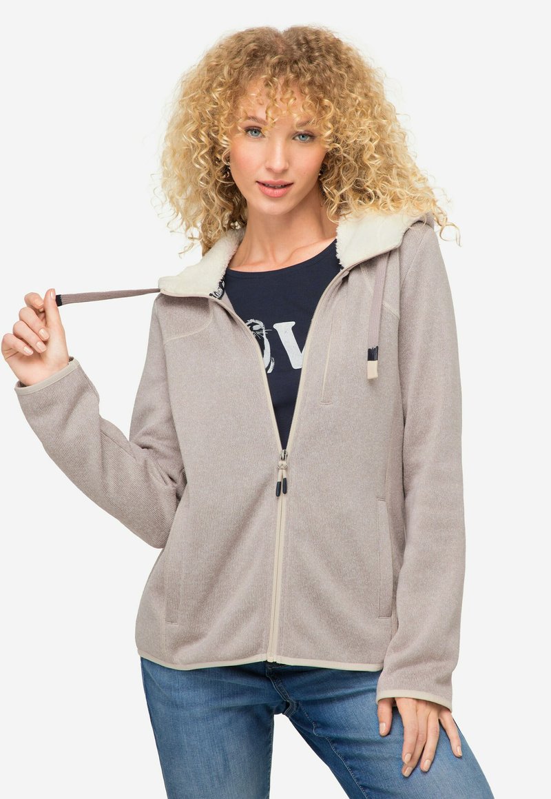 Sudadera con cremallera de color beige claro, con collar forrado de felpa, dos bolsillos frontales y textura acanalada. Tiene una cremallera de contraste y capucha con cordón.