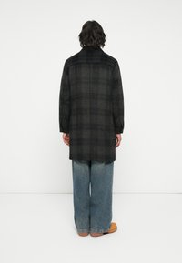 Sort og grå ternet lang frakke med lige snit, båret over løse blå jeans. Stoffet ser varmt og tekstureret ud med et klassisk design.