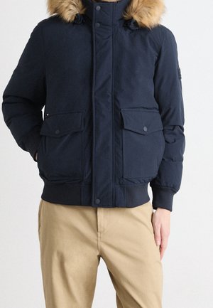 Marineblaue gepolsterte Jacke mit fellgefütterter Kapuze, Druckknöpfen, zwei Fronttaschen und gerippten Bündchen. Glatte Stoffoberfläche.