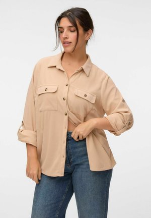 Femme aux cheveux foncés portant une chemise beige boutonnée avec les manches retroussées et un jean bleu, tirant légèrement la chemise au niveau de la taille.