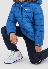 Blå puffjacka med dragkedja fram och huva. Bärs med marinblå joggingbyxor. Vita sneakers med blå sulor och röda detaljer.