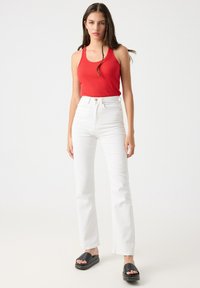 Top sin mangas de rayas rojas combinado con jeans blancos de talle alto. La modelo lleva sandalias de plataforma negras con detalles de tiras. Fondo sencillo.