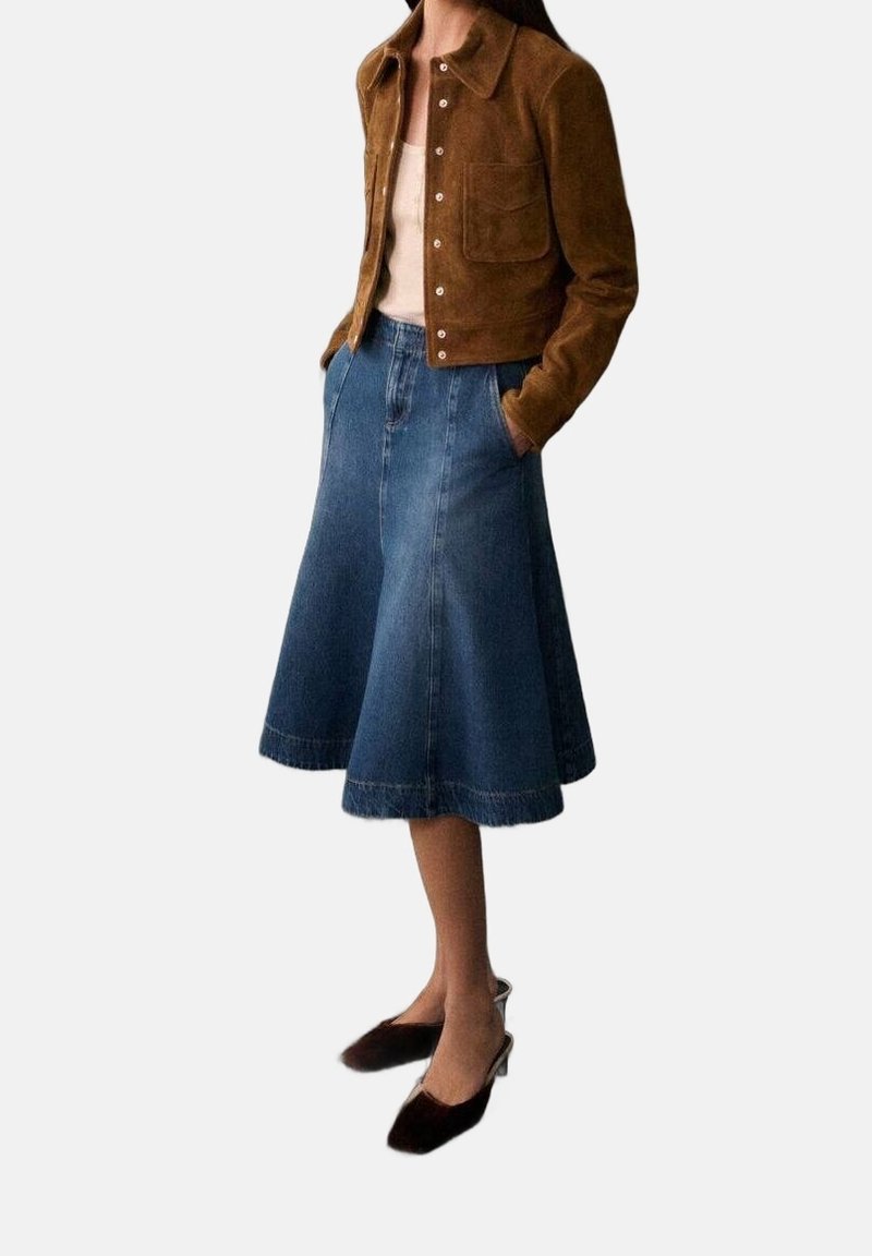Femme portant une veste en daim marron, un haut beige, une jupe en denim évasée longueur genou et des chaussures slingback à bouts pointus foncées, debout avec les mains dans les poches.