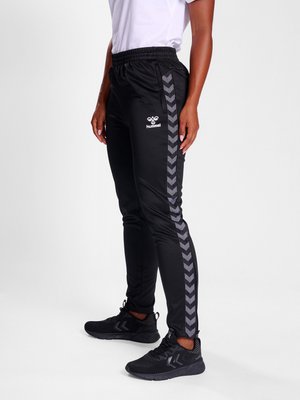 Person iført sorte Hummel træningsbukser med grå chevronstriber ned langs siderne, sorte sneakers og en hvid skjorte, stående sidelæns.